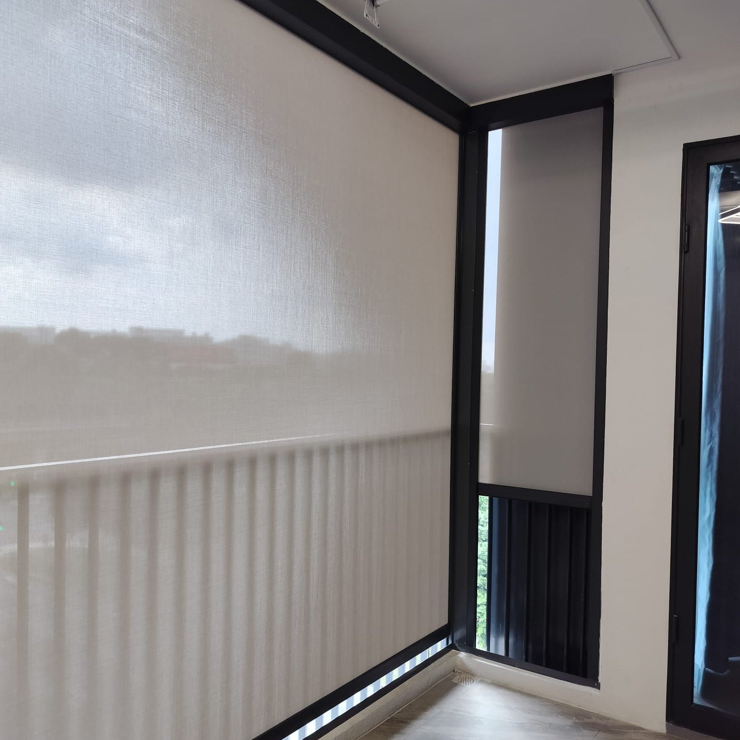 A large white roller blind with a black frame installed on a balcony or terrace wall, with a matching blind partially drawn on an adjacent door. The room has white walls, a gray floor, and ceiling fixtures visible.