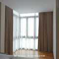 A pair of floor-length taupe brown pleated curtains paired with sheer white window drapes, installed on a bay window in a modern room with light wood flooring.