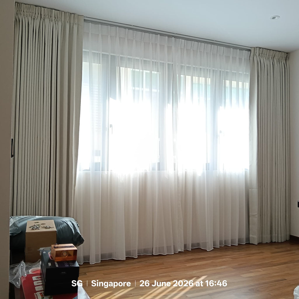 A set of beige pleated window curtains and a white sheer drape hanging over a large window in a room with hardwood floors. A cardboard box and other items are visible on the floor in the lower left corner.