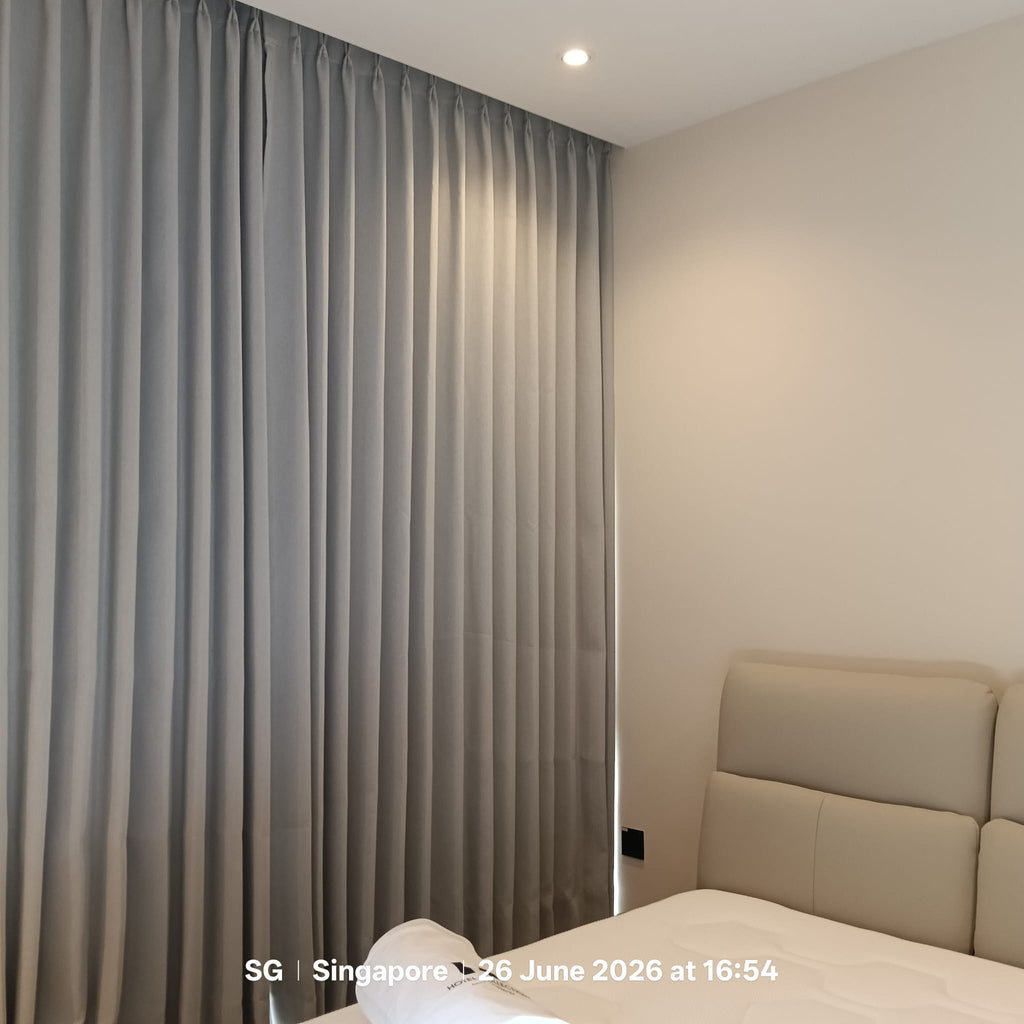 A pair of floor-length gray pleated curtains hanging in a bedroom corner next to a bed with a beige upholstered headboard.